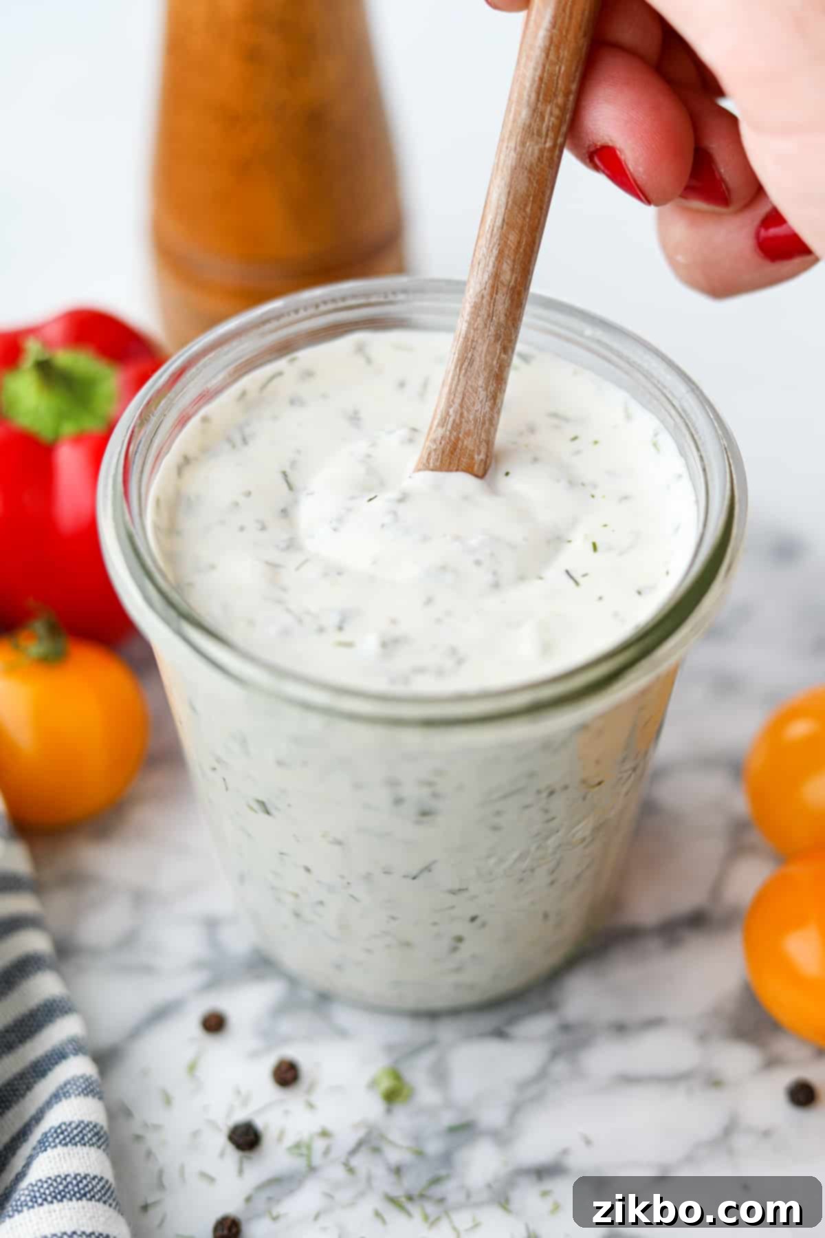 Zesty Greek Ranch Duo 9 A hand stirring creamy homemade Greek Yogurt Ranch in a glass jar, highlighting its smooth, consistent texture and vibrant herb flecks.