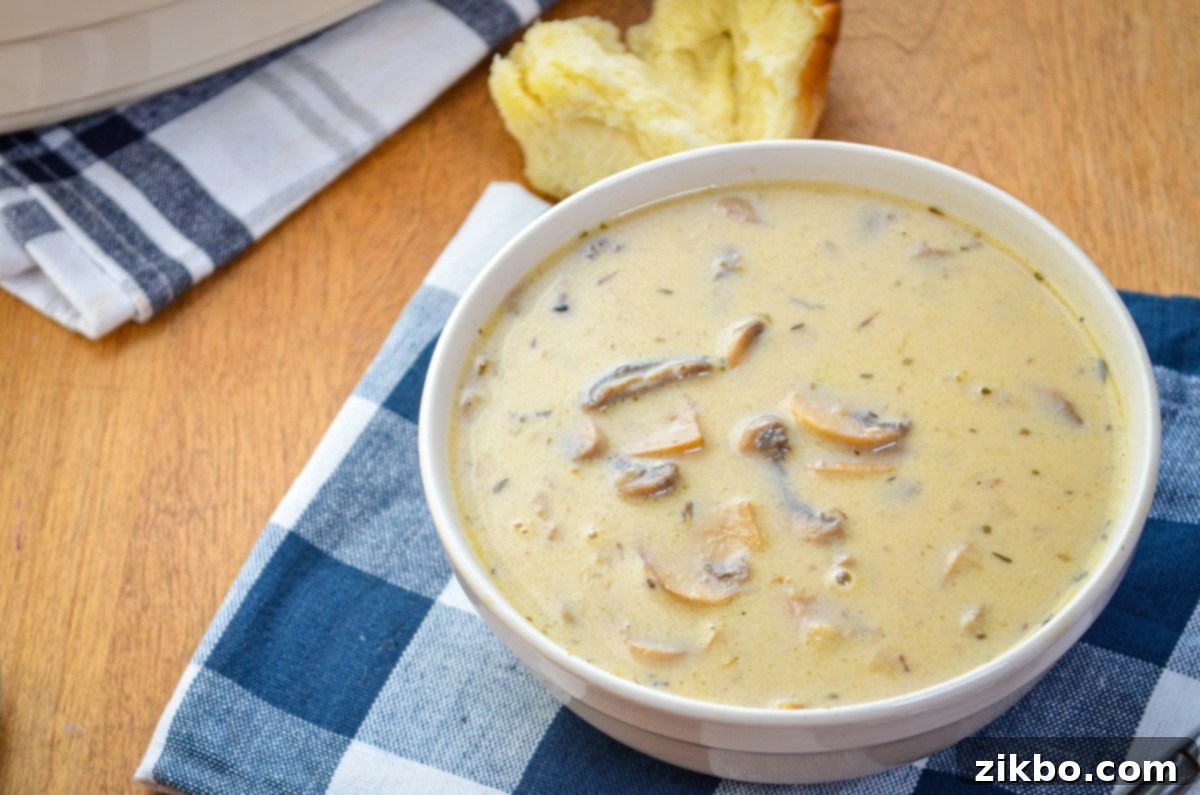 A bowl of cream of mushroom soup with a torn roll in the background.