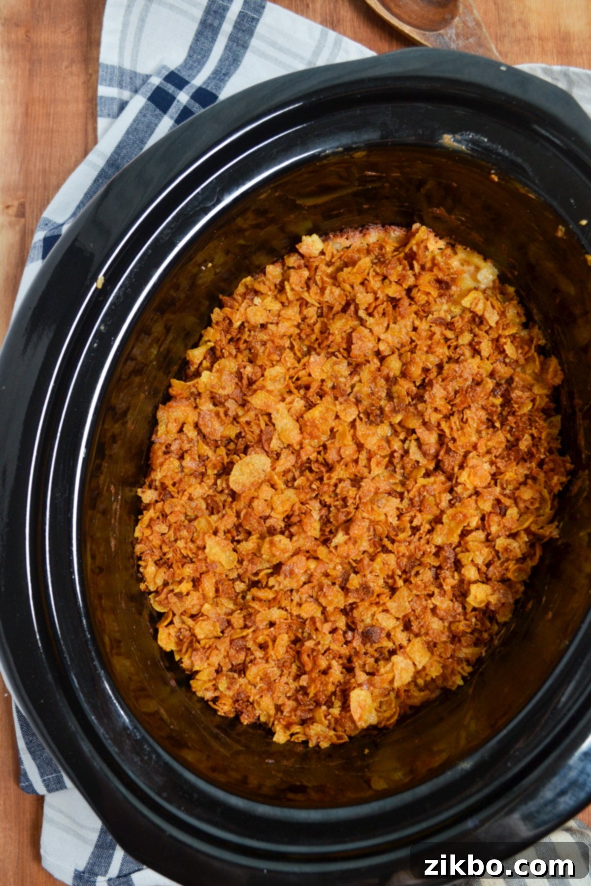 Slow Cooker Golden Cheesy Hash Brown Casserole 10 A crock pot containing a rich, cheesy hash brown casserole, topped with a golden and crunchy cornflake crust, resting elegantly on a decorative napkin.