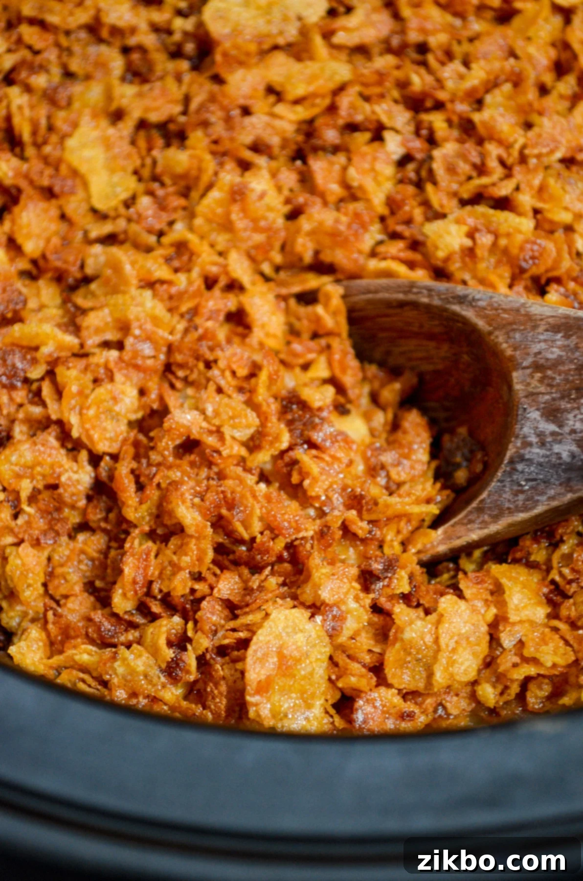 Slow Cooker Golden Cheesy Hash Brown Casserole 4 A wooden spoon scoops a generous portion of cheesy hash brown casserole, crowned with a golden, crispy cornflake topping, directly from a slow cooker.
