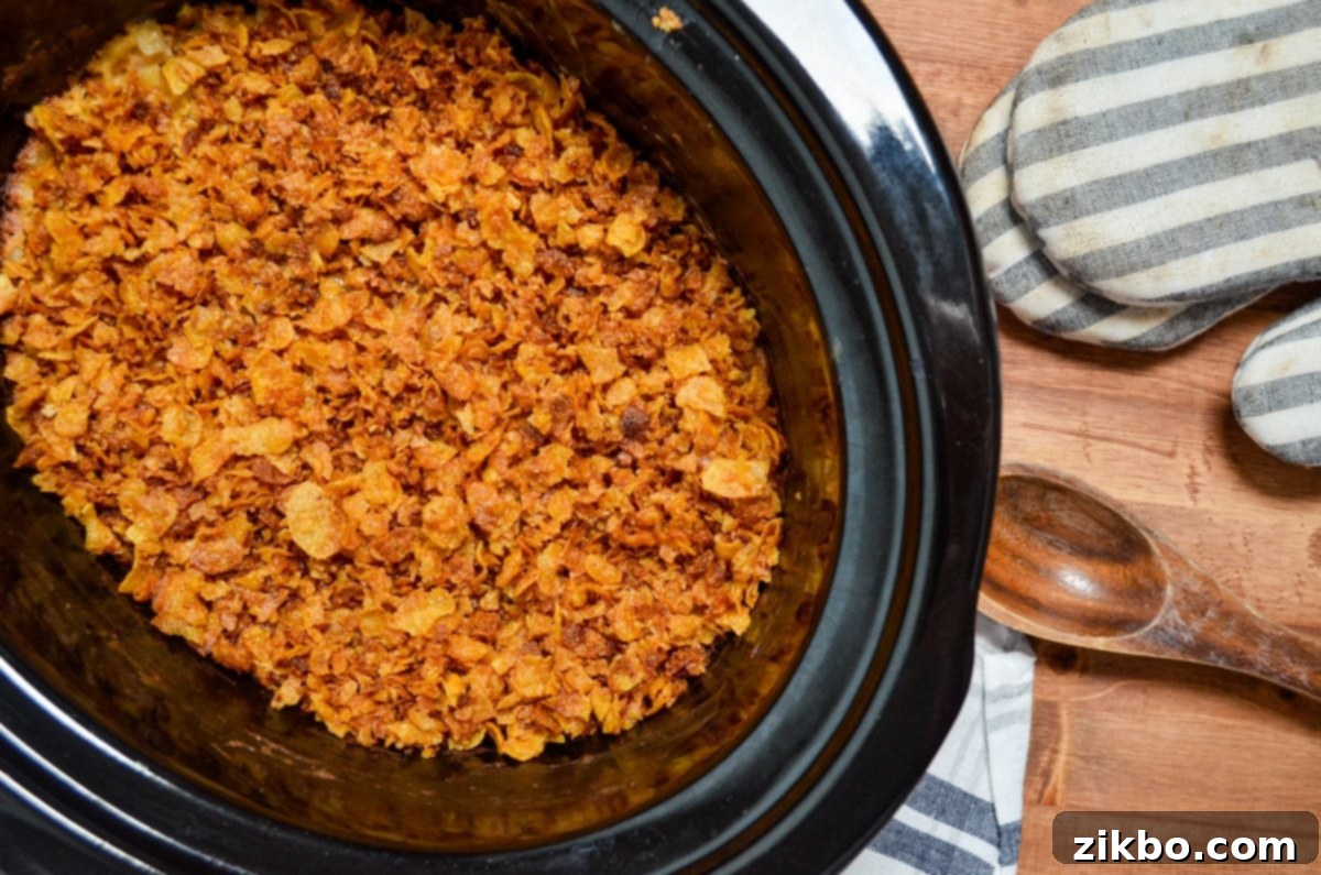 Slow Cooker Golden Cheesy Hash Brown Casserole 9 A large crock pot full of cheesy hash brown casserole, adorned with a perfectly crispy crushed corn flake topping, resting on a napkin with a wooden spoon and oven mitts beside it.