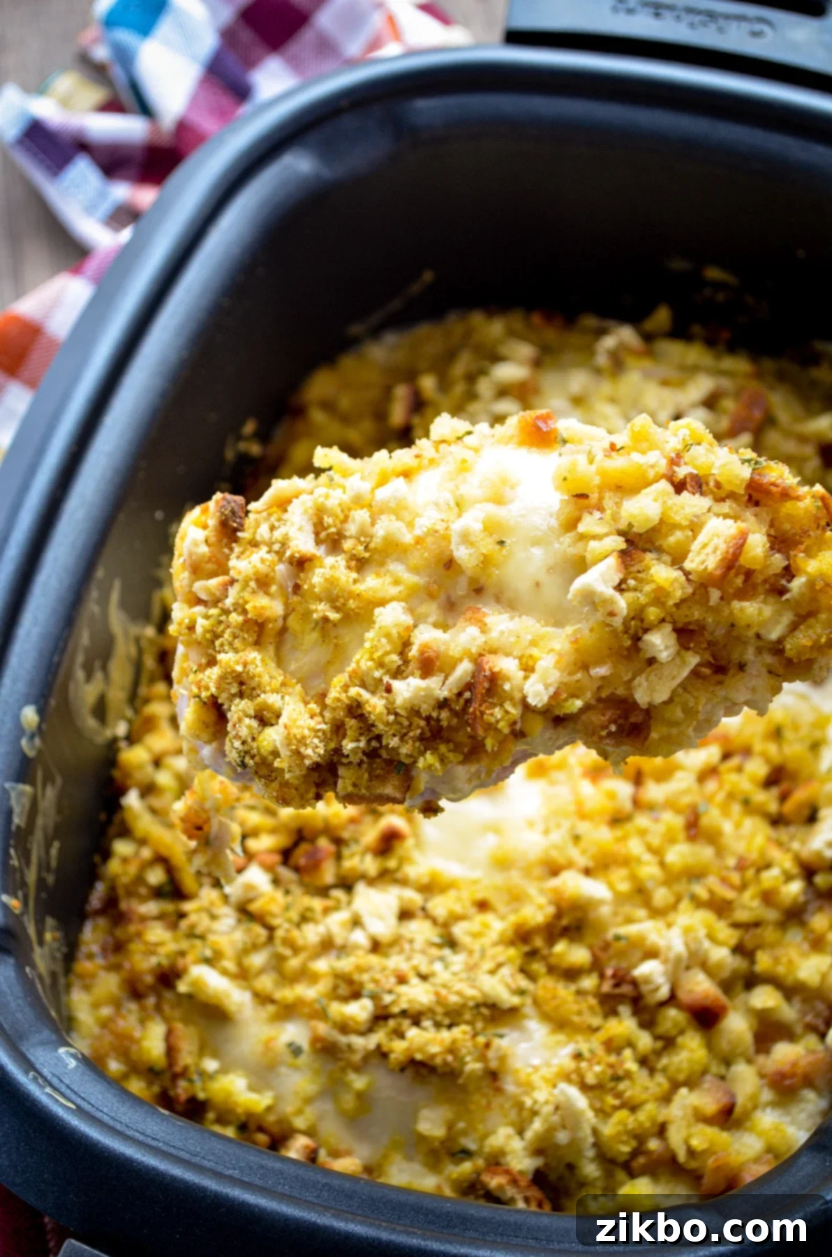 A wooden spoon lifting out a portion of Swiss Chicken Casserole from a Slow Cooker. A chicken breast covered in swiss cheese, a simple sauce, and stove top stuffing.