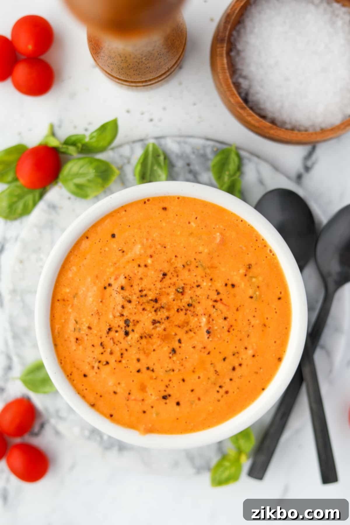 A bowl of creamy tomato soup, surrounded by fresh basil leaves.