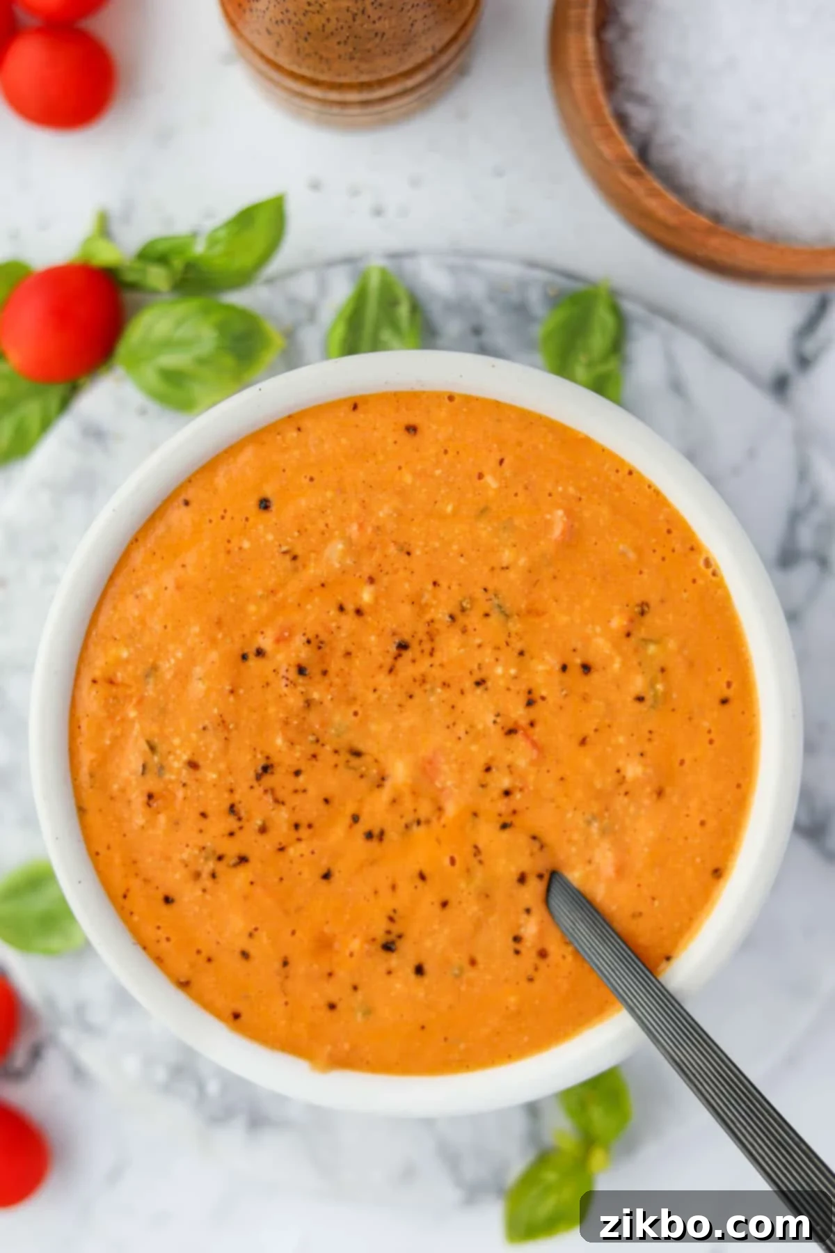 An overhead view of a bowl of tomato soup, with basil and cherry tomatoes around the counter.