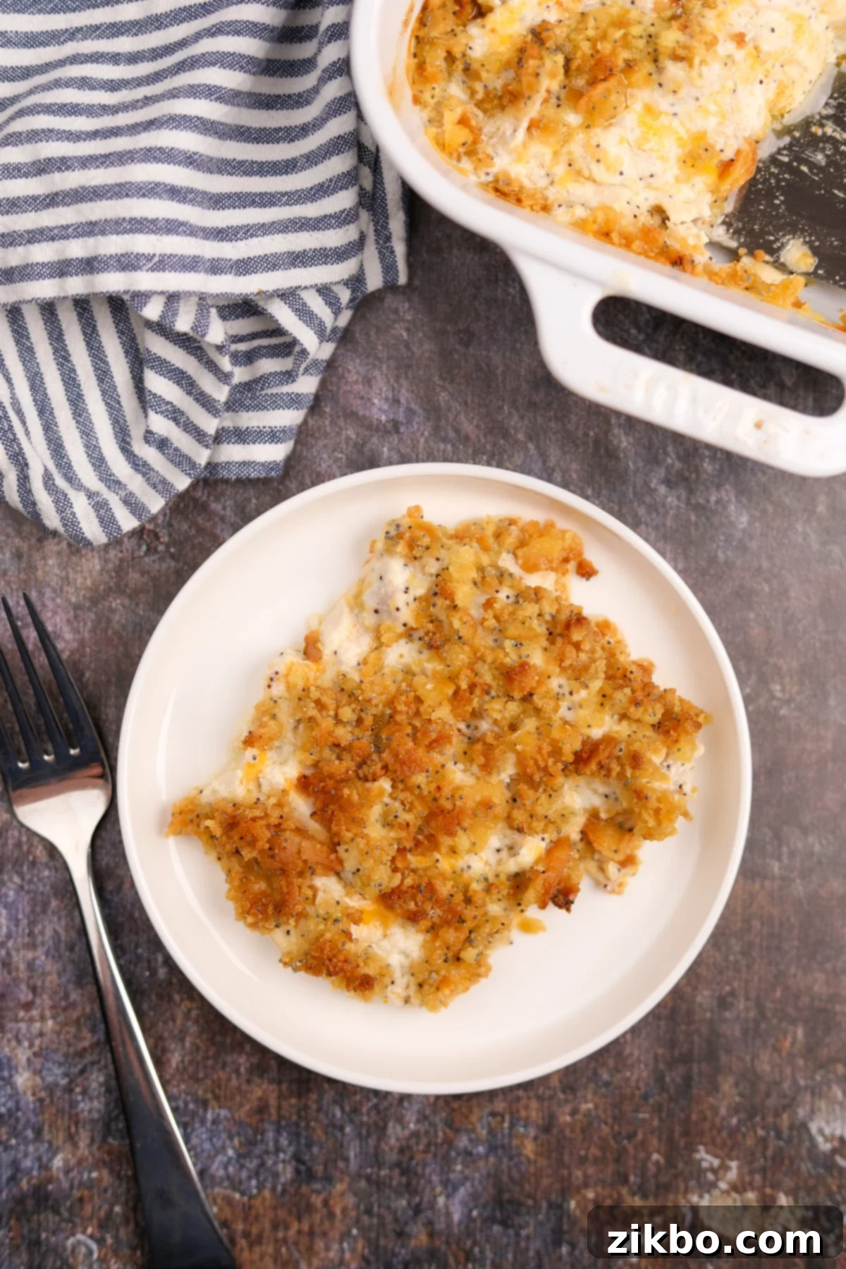 A beautifully plated portion of gluten-free poppy seed chicken casserole on a white plate, with the larger baking dish visible in the background, highlighting the creamy texture and crunchy topping.