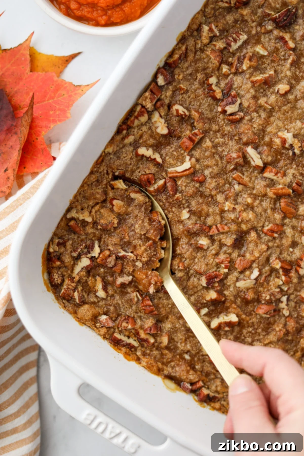 A pecan streusel topped pumpkin casserole in a white dish, with a gold serving spoon taking a portion.
