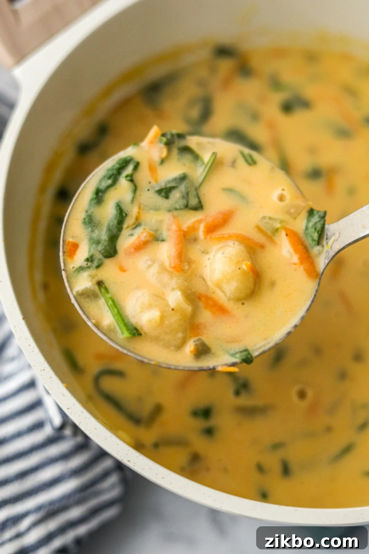 A ladleful of cheesy gnocchi soup, showing the potato dumplings, spinach, and shredded carrots.