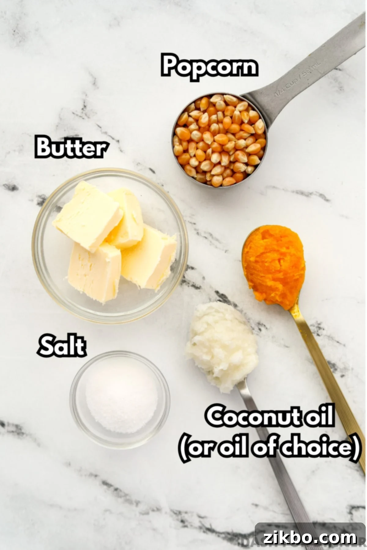 The ingredients needed to make stovetop popcorn: popcorn kernels, a bottle of coconut oil, a stick of butter, and a small dish of salt, laid out on a clean surface.