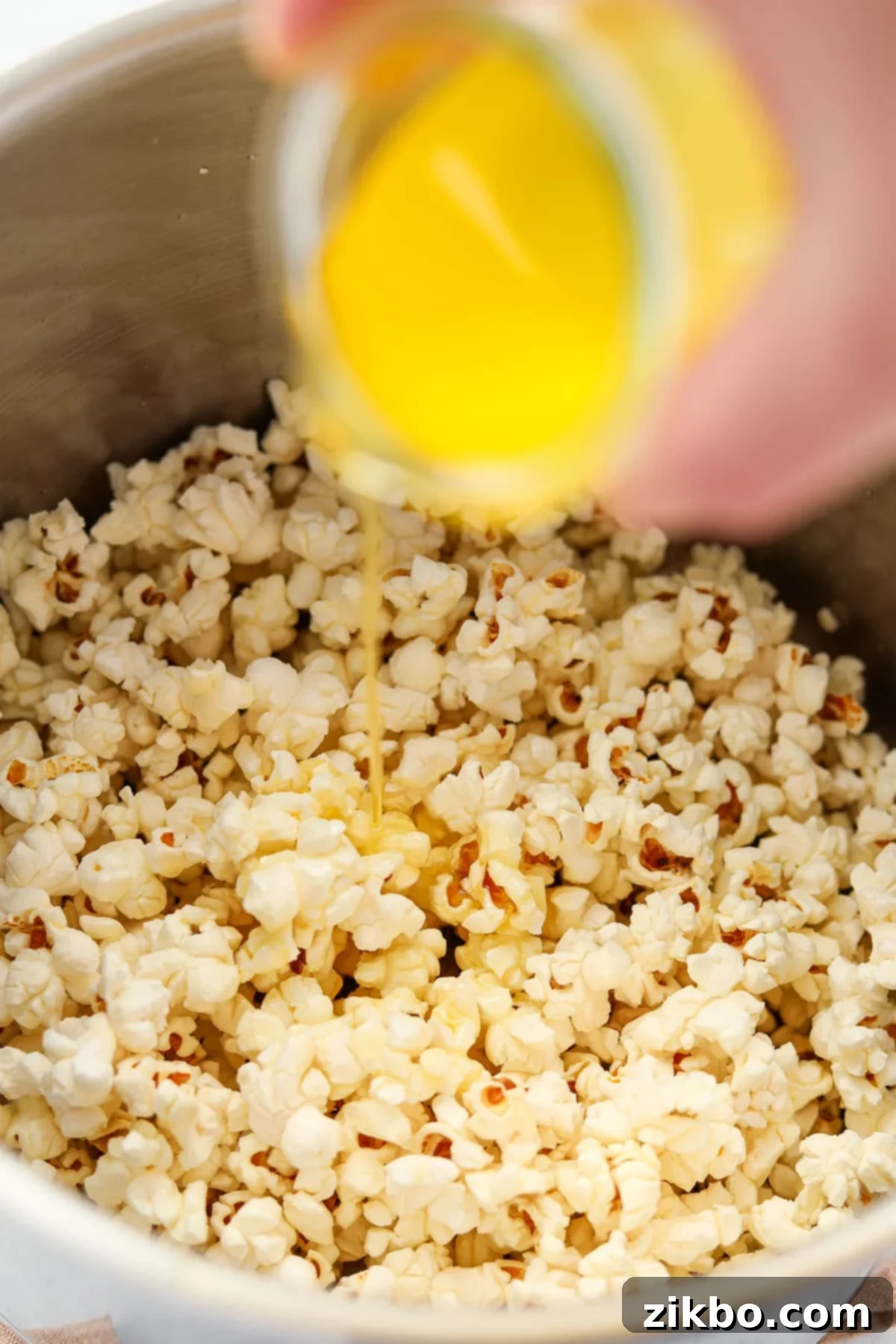 Drizzling melted butter over popped kernels in a large pot, emphasizing even coating for delicious popcorn.