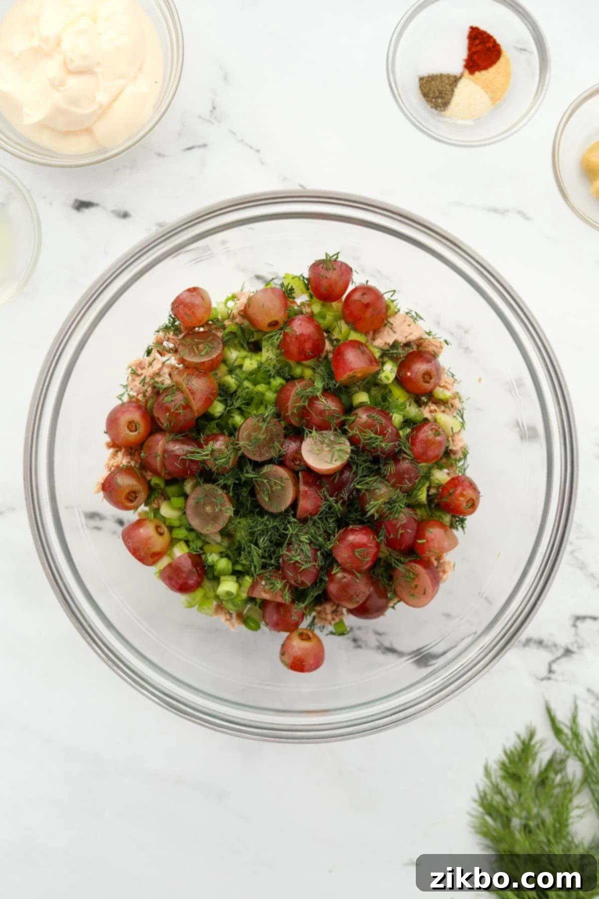 A bowl with drained tuna, halved grapes, finely diced celery, chopped green onion, and fresh dill, ready to be mixed.