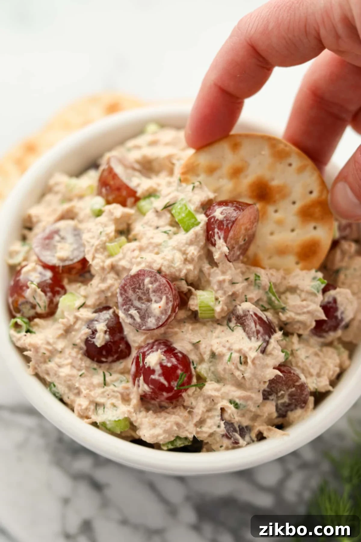 Dipping a cracker into a bowl of tuna salad with grapes, showing the creamy texture and visible pieces of tuna and grape.