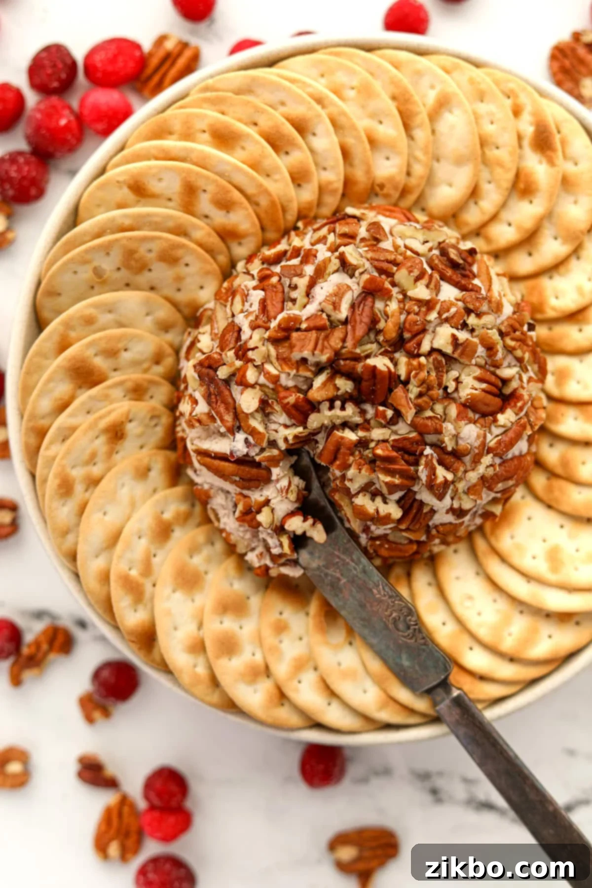 An appetizer knife taking a portion of a creamy Boursin cheese ball coated in chopped pecans from a festive serving platter.