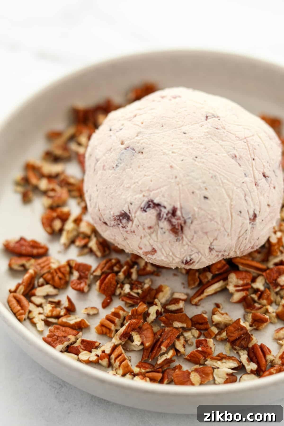 A chilled Boursin cheese ball being rolled in a shallow dish filled with finely chopped pecans, ensuring full coverage.