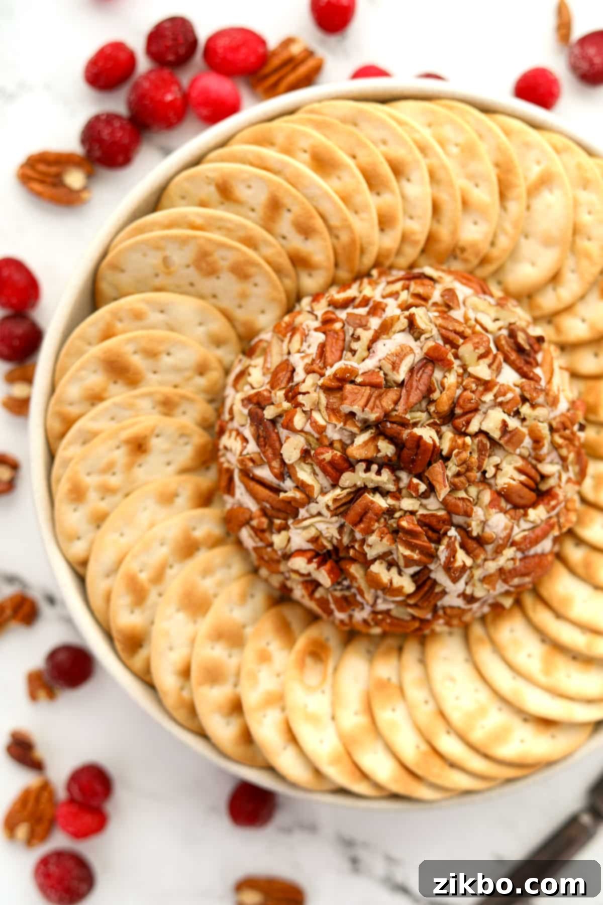 A beautifully presented pecan-crusted Boursin cheese ball, surrounded by an assortment of crackers and an appetizer knife on a white platter.