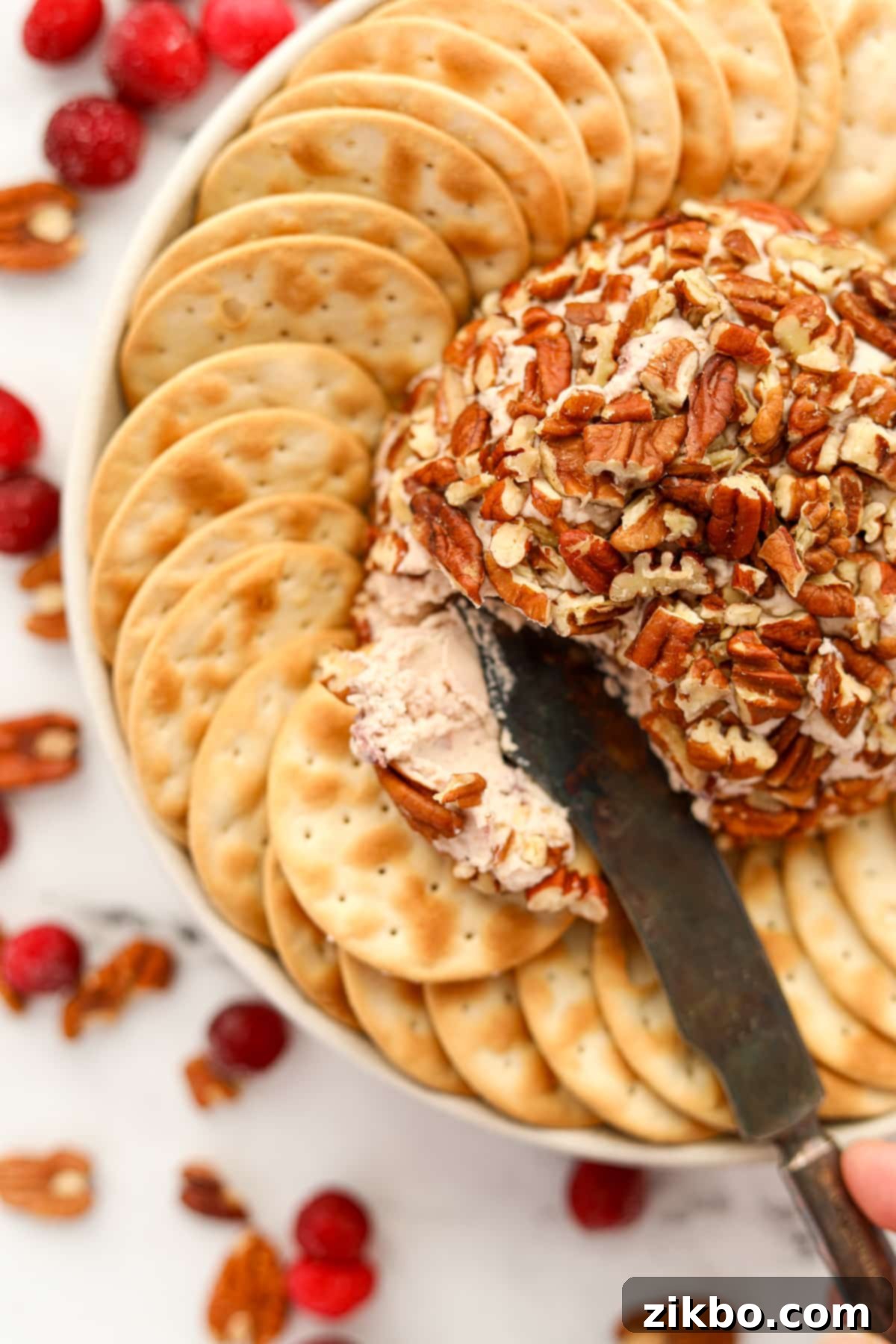 A close-up of an appetizer knife scooping a generous portion of creamy Boursin cheese from a pecan-crusted cheese ball, ready to be spread.
