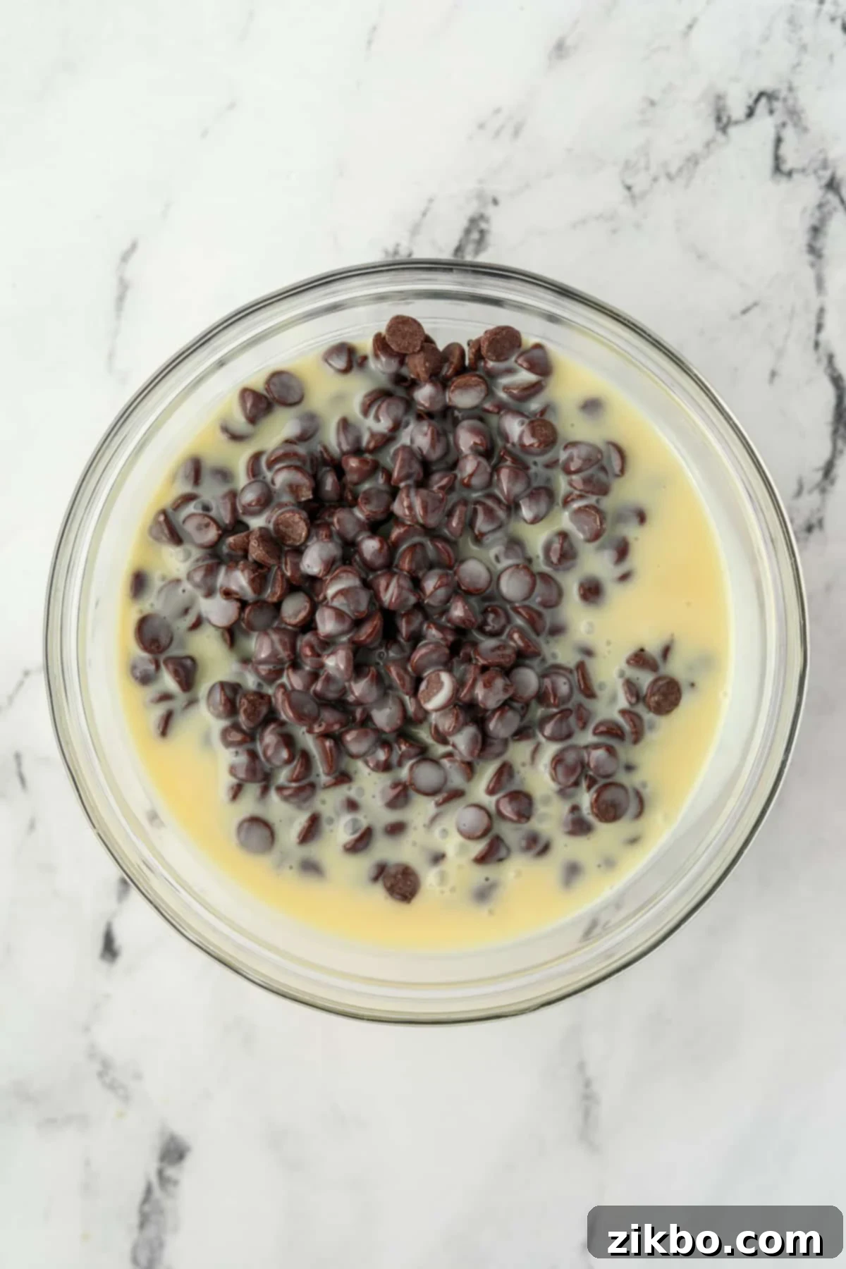 A large glass bowl filled with chocolate chips and sweetened condensed milk, ready for microwaving.