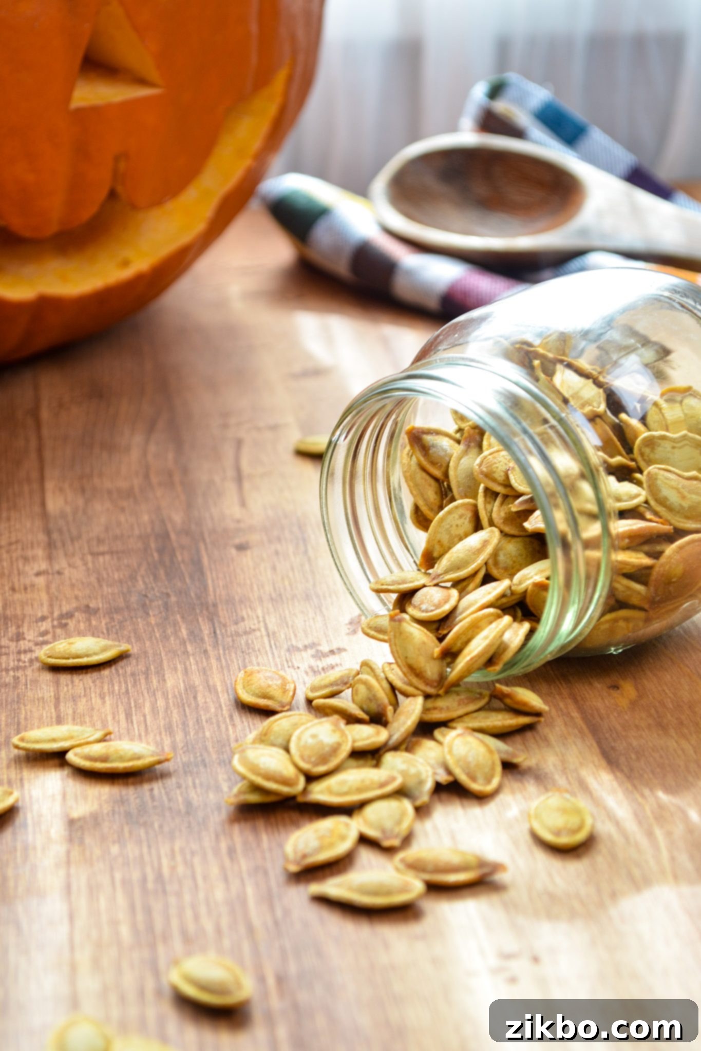 A jar of roasted pumpkin seeds being spilled onto a rustic wood table, with a festive pumpkin in the background.