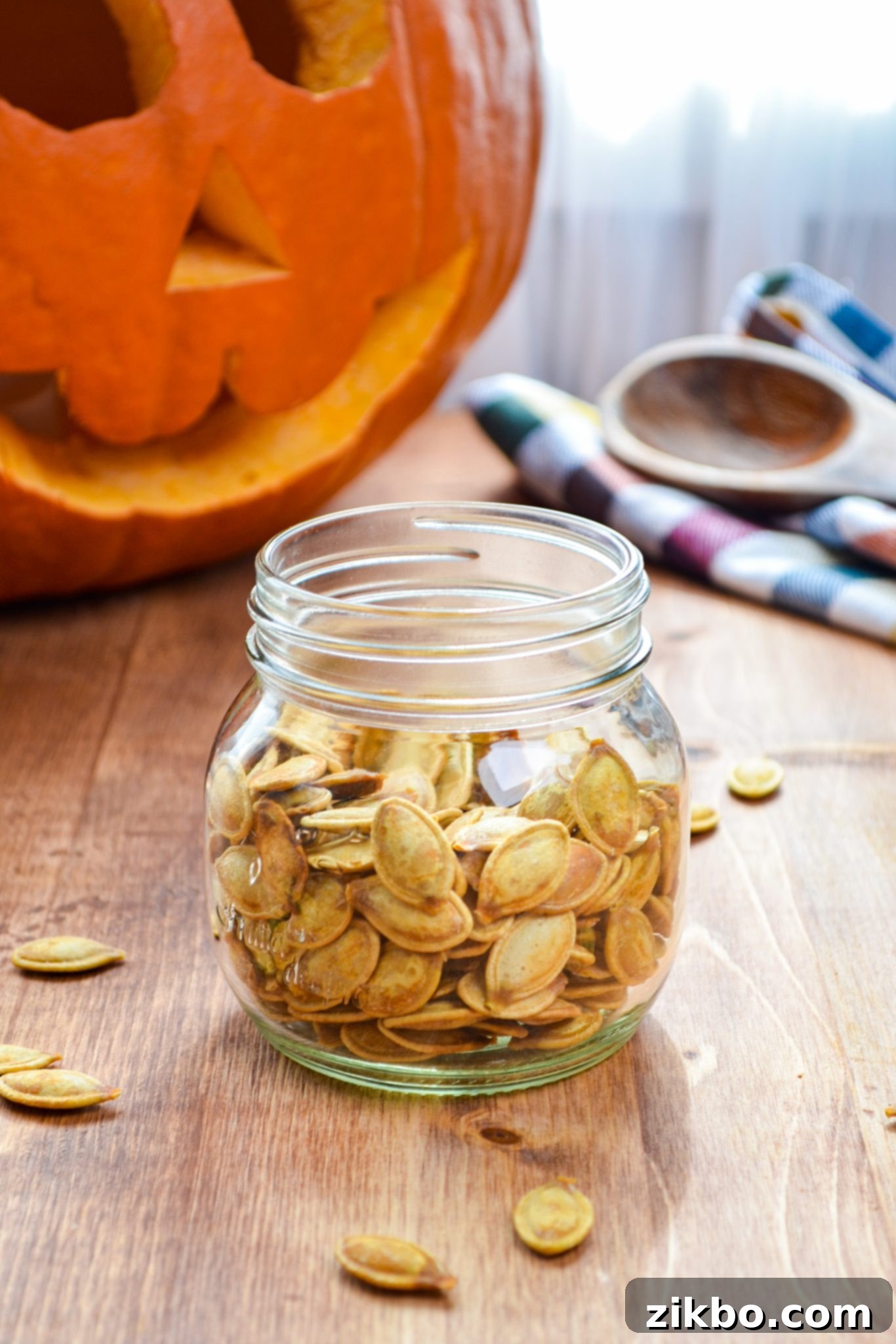 A jar of roasted pumpkin seeds, with a large carved Halloween pumpkin blurred in the background, setting a festive fall scene.