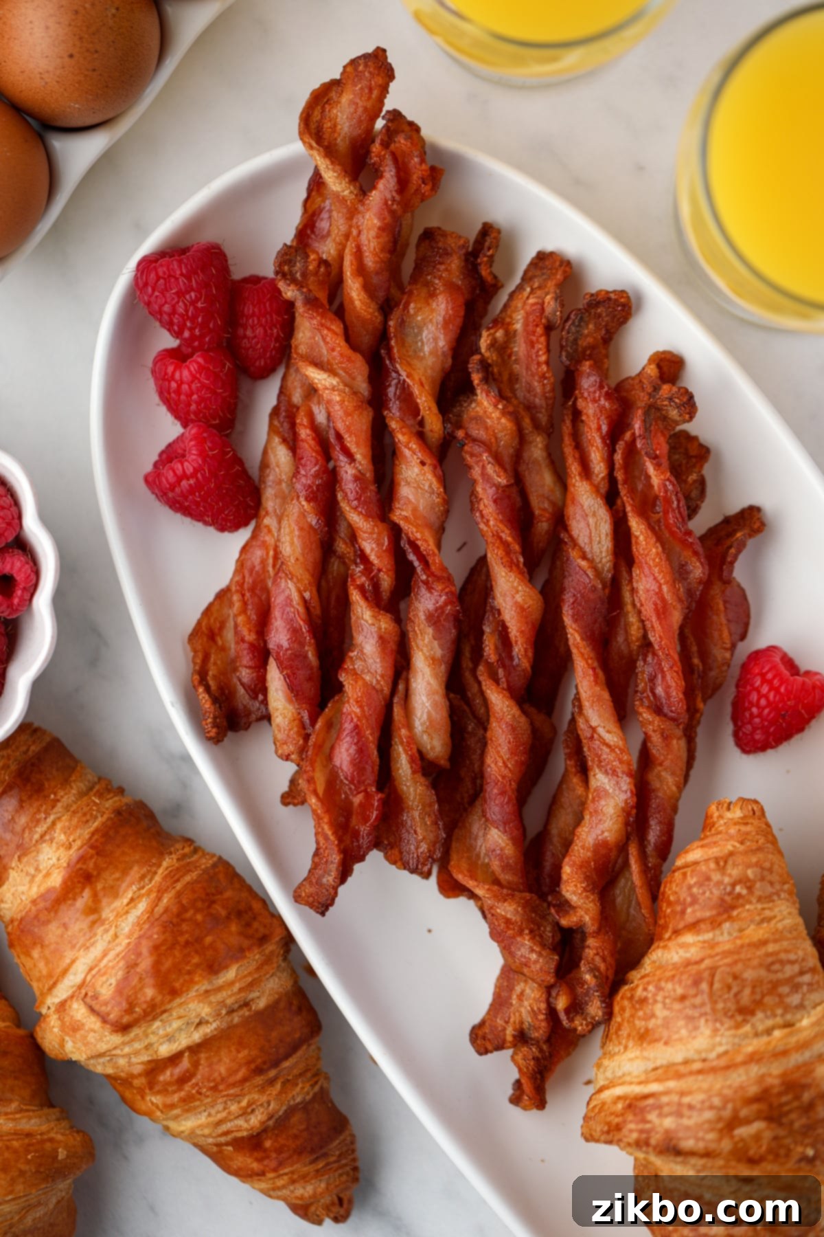 Crispy twisted bacon on a white platter, garnished with fresh raspberries and some croissants on the side.