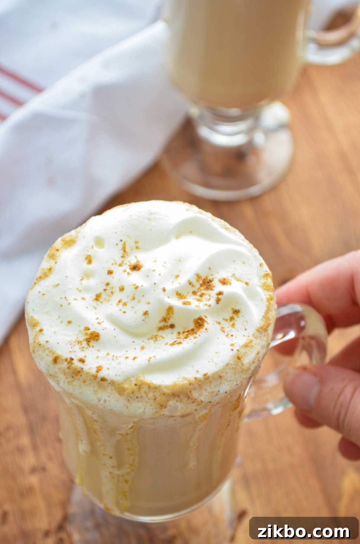 A hand gently lifting a clear glass mug of an Instant Pot Pumpkin Spice Latte, showcasing the rich coffee color and the inviting swirl of whipped cream on top, ready for sipping.