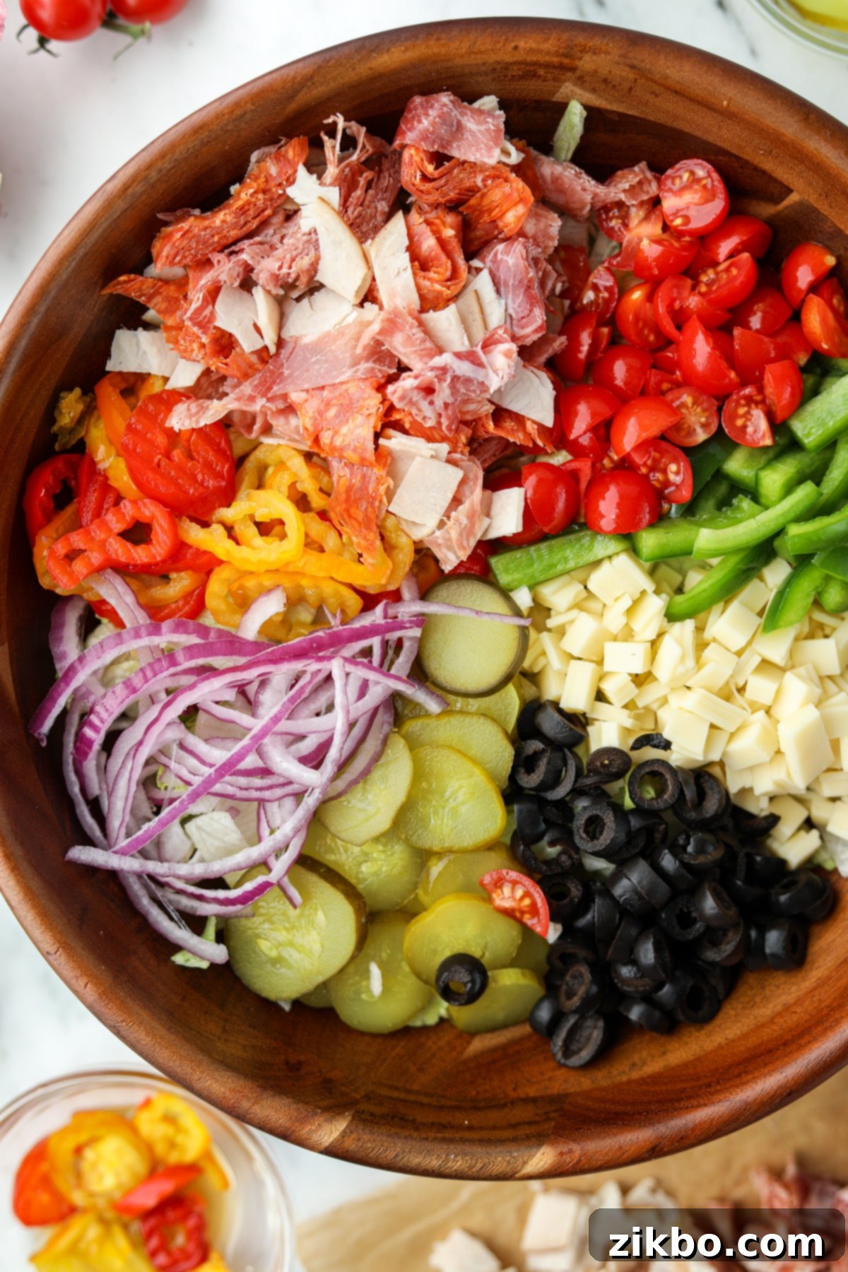 An overhead image of a large salad bowl filled with beautifully arranged sections of toppings, including deli meat, cheese, pickles, and other vegetables, before being tossed.