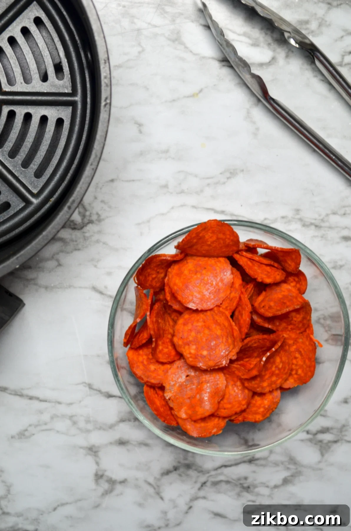 A bowl of pepperoni, with an air fryer basket and a pair of tongs on the side.