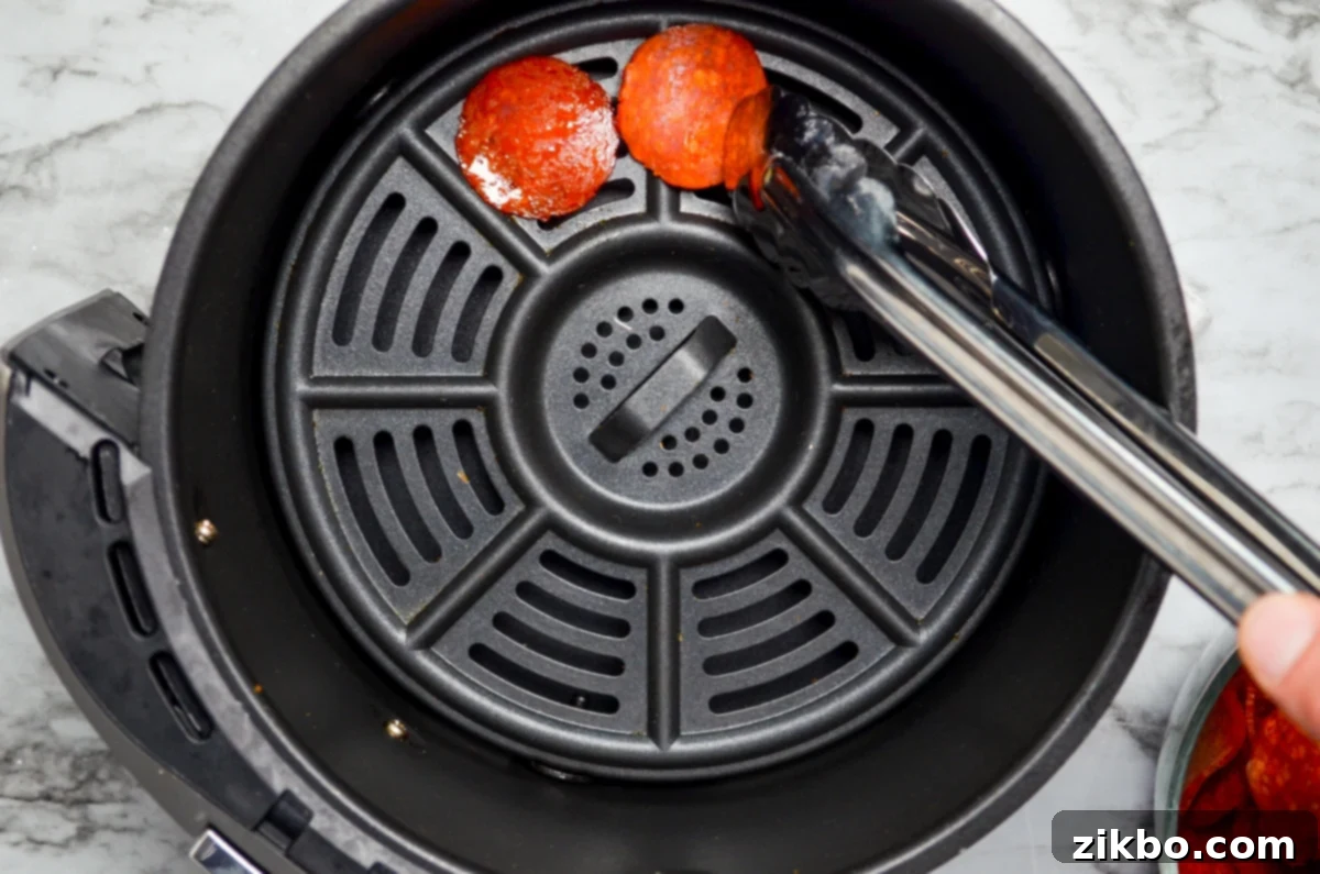 A set of tongs placing pepperoni slices in the basket of an air fryer.