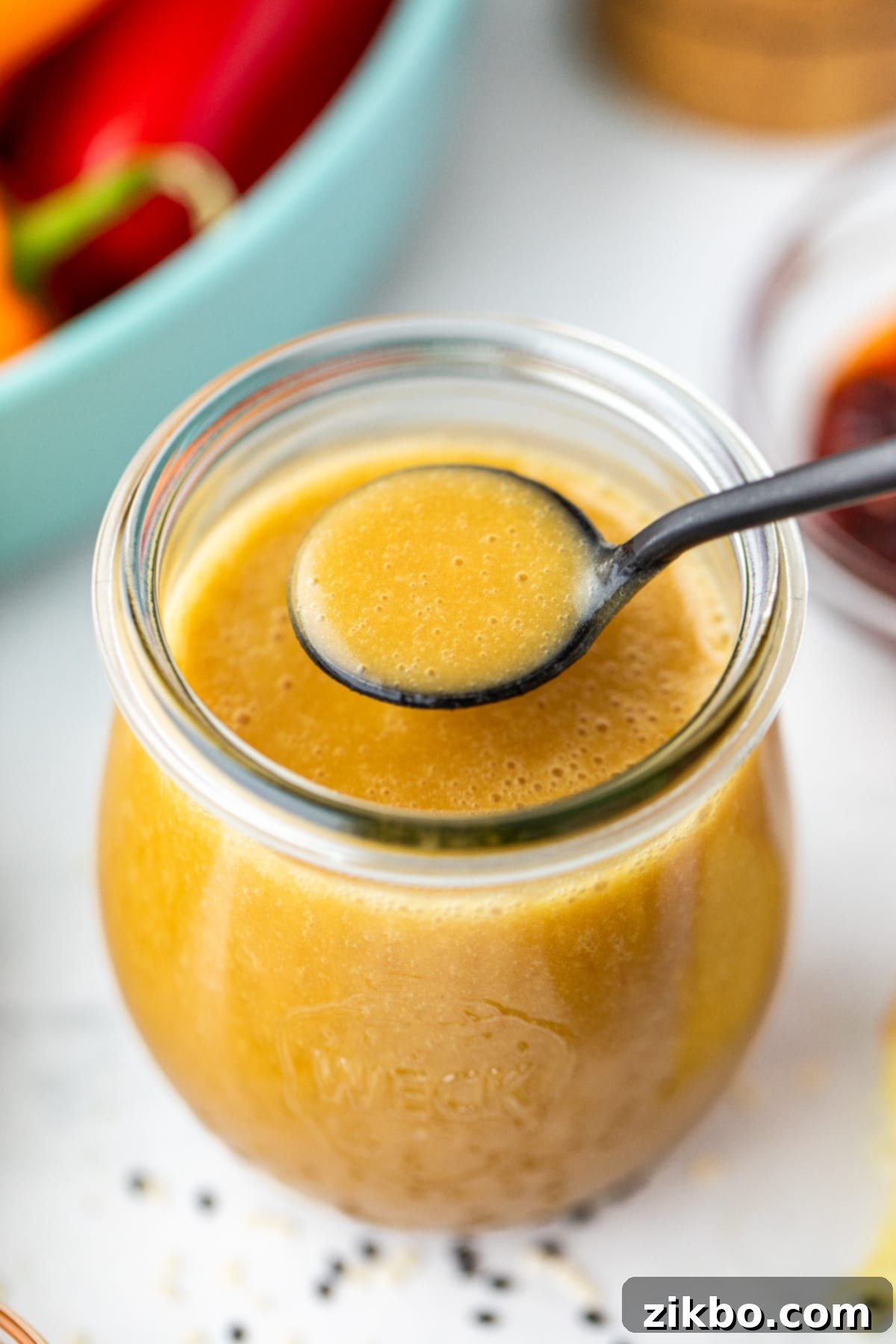 Sesame Ginger Dressing 8 A spoon delicately taking a small amount of the golden homemade vinaigrette from a clear glass jar, showcasing its smooth texture.