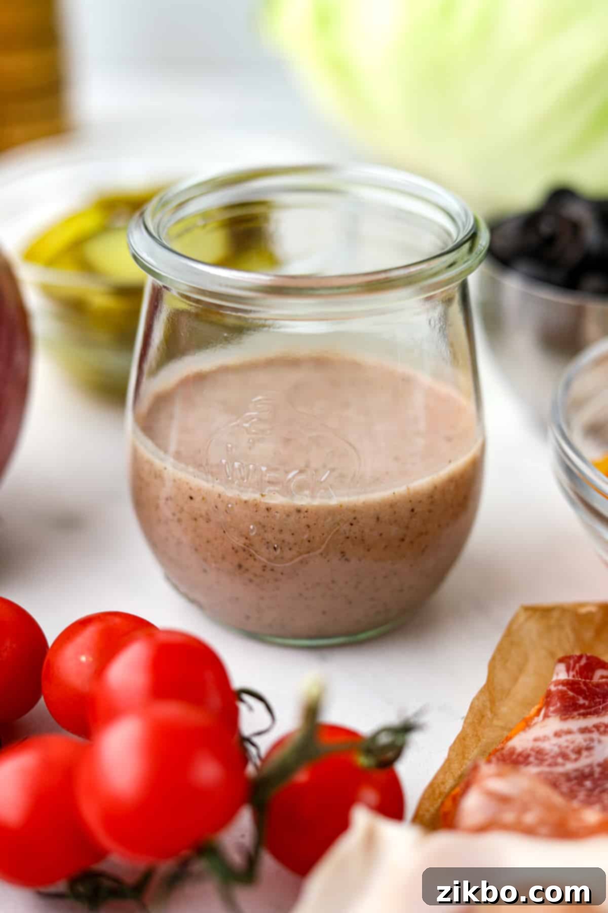 A beautifully presented small glass jar filled with vibrant homemade Italian sub dressing, artfully surrounded by an assortment of fresh ingredients including cherry tomatoes, crisp lettuce, black olives, and thinly sliced cured meats, highlighting its versatile applications.