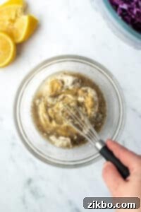 A hand whisking together mayonnaise, lemon juice, dijon mustard, and black pepper in a small bowl to create a smooth, creamy dressing.