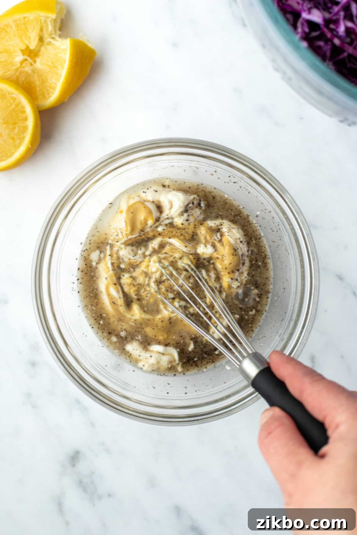 Zesty Israeli Cabbage Slaw 7 A hand whisking together mayonnaise, lemon juice, dijon mustard, and black pepper in a bowl to create a smooth, creamy dressing.
