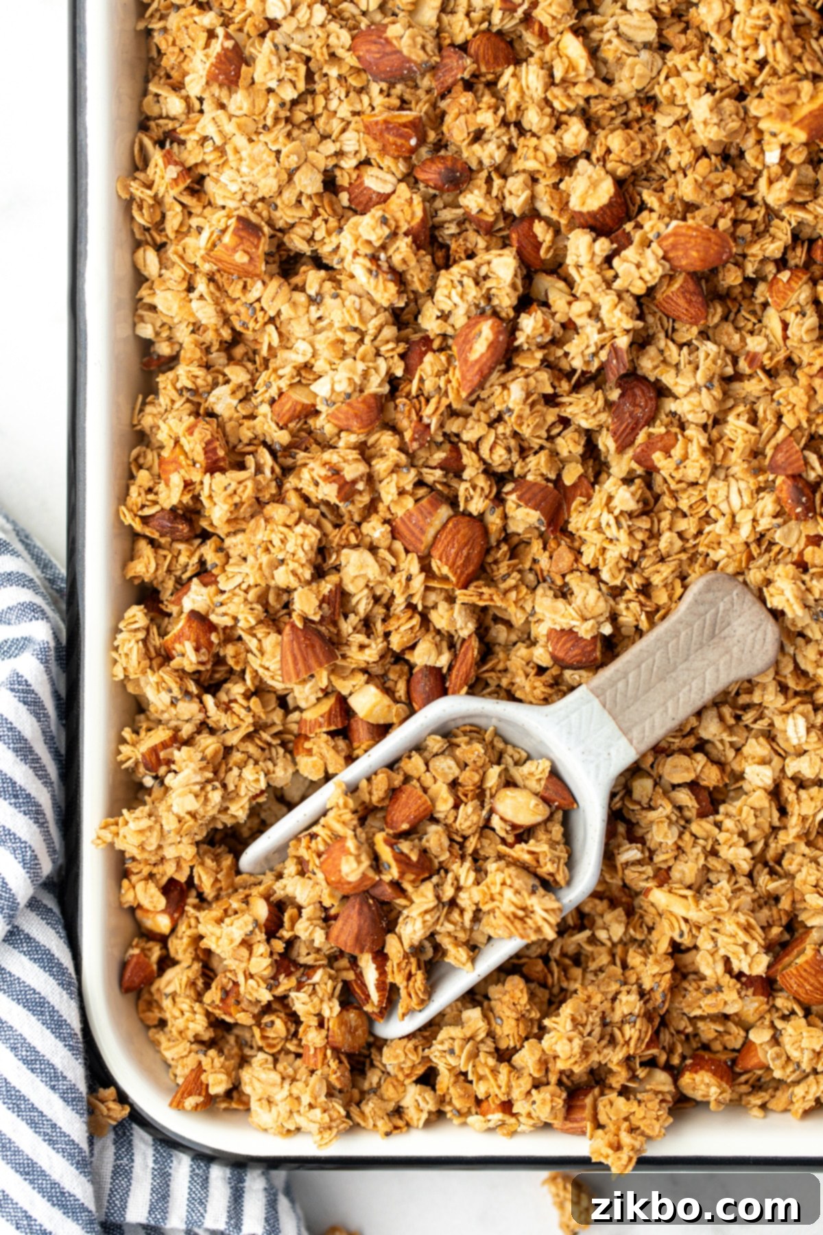 A baking sheet filled with golden-brown granola, with a scoop taking a small portion, highlighting its crunchy texture.