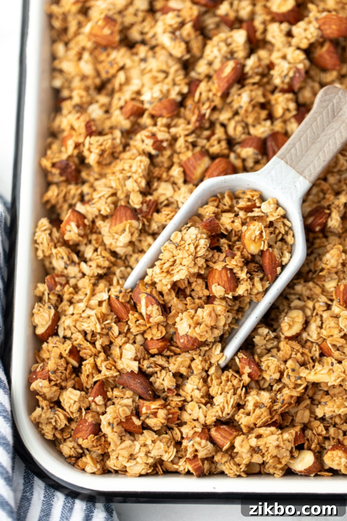 A scoop taking a portion of golden-brown granola from a sheet pan, showcasing its clumped texture.