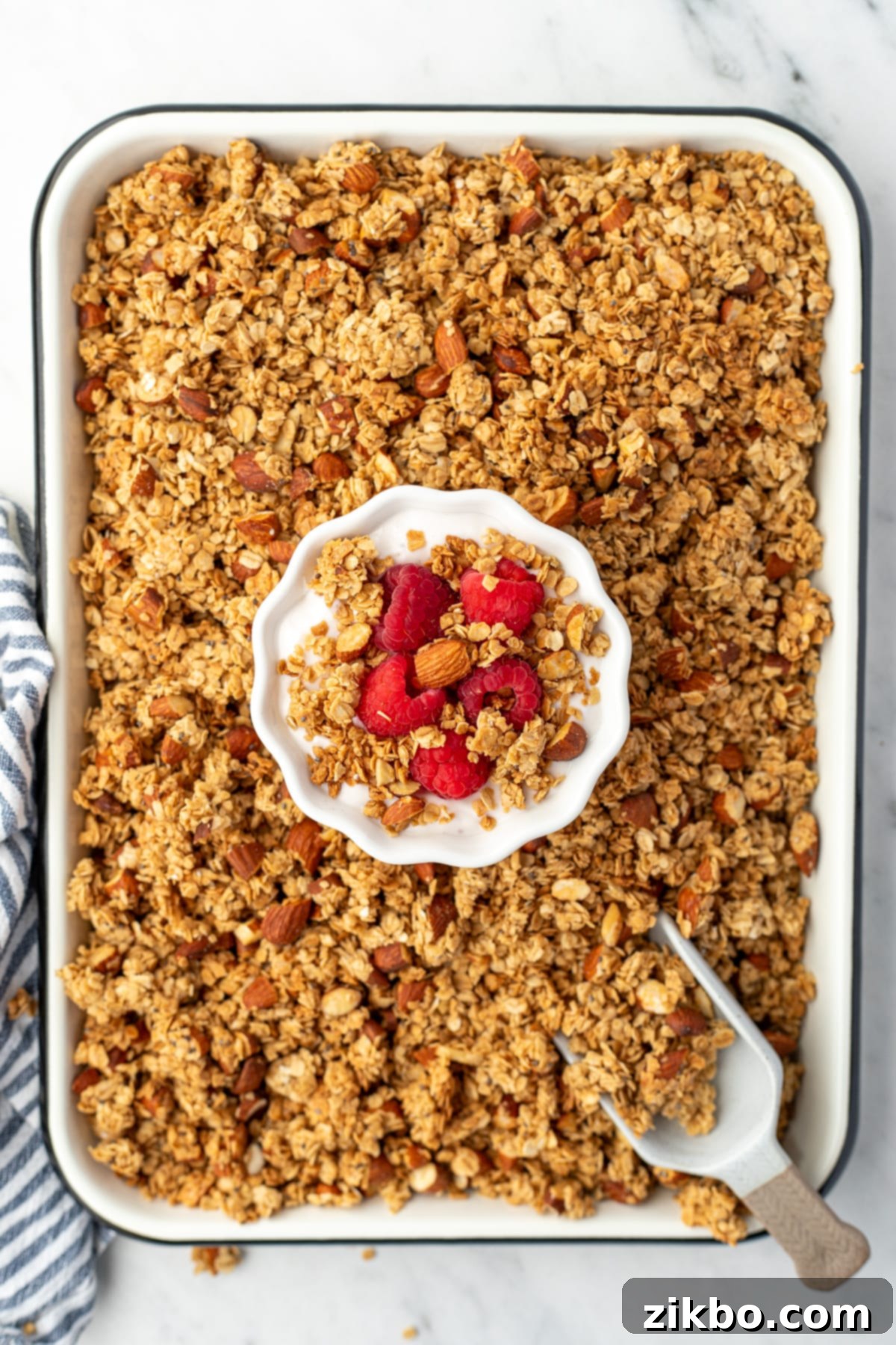 A sheet pan filled with baked golden-brown granola, with a small bowl of creamy yogurt and fresh berries nestled in the center.