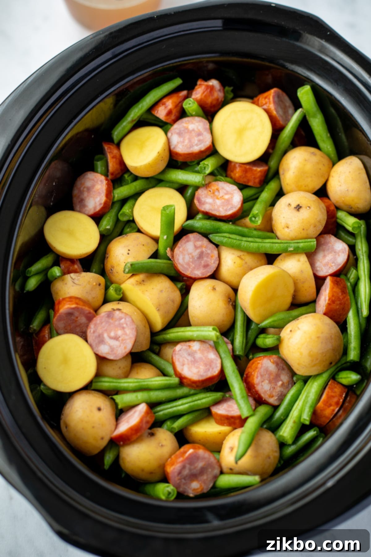 A slow cooker filled with vibrant baby potatoes, sliced smoked sausage, and fresh green beans, ready for seasoning.