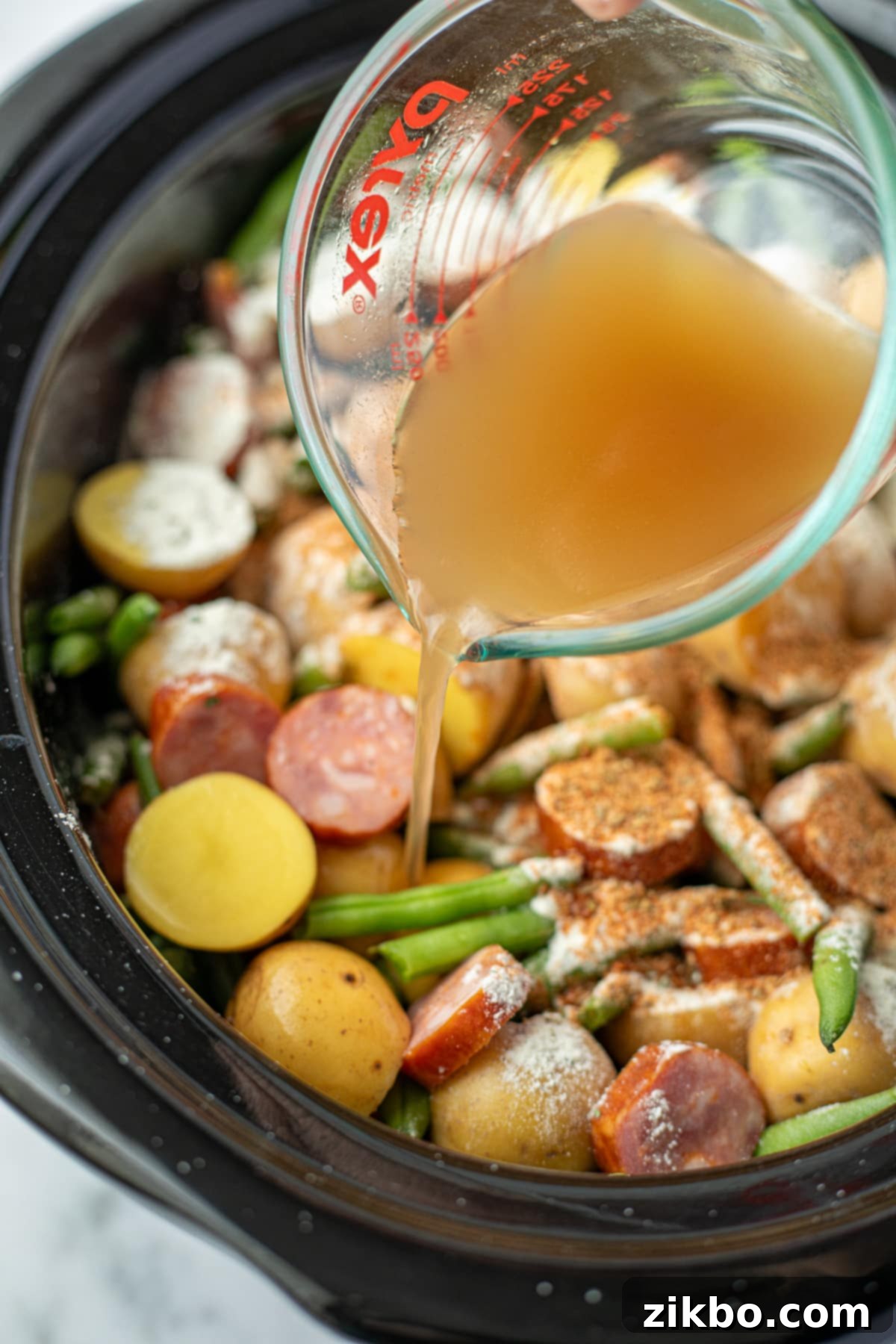 A stream of rich beef broth being poured into a slow cooker over potatoes, green beans, and smoked sausage, preparing for slow cooking.