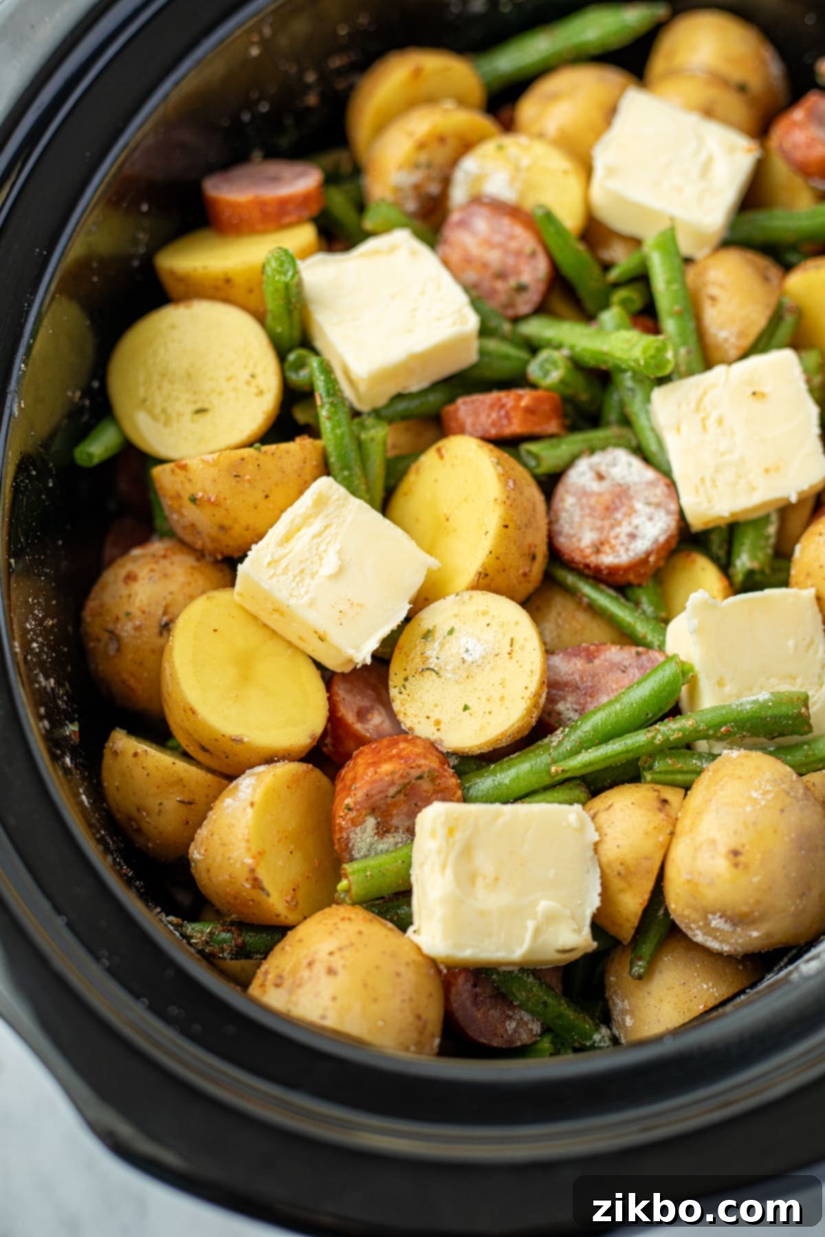 A slow cooker filled with potatoes, green beans, and smoked sausage, now topped with six evenly spaced slices of butter, ready for cooking.