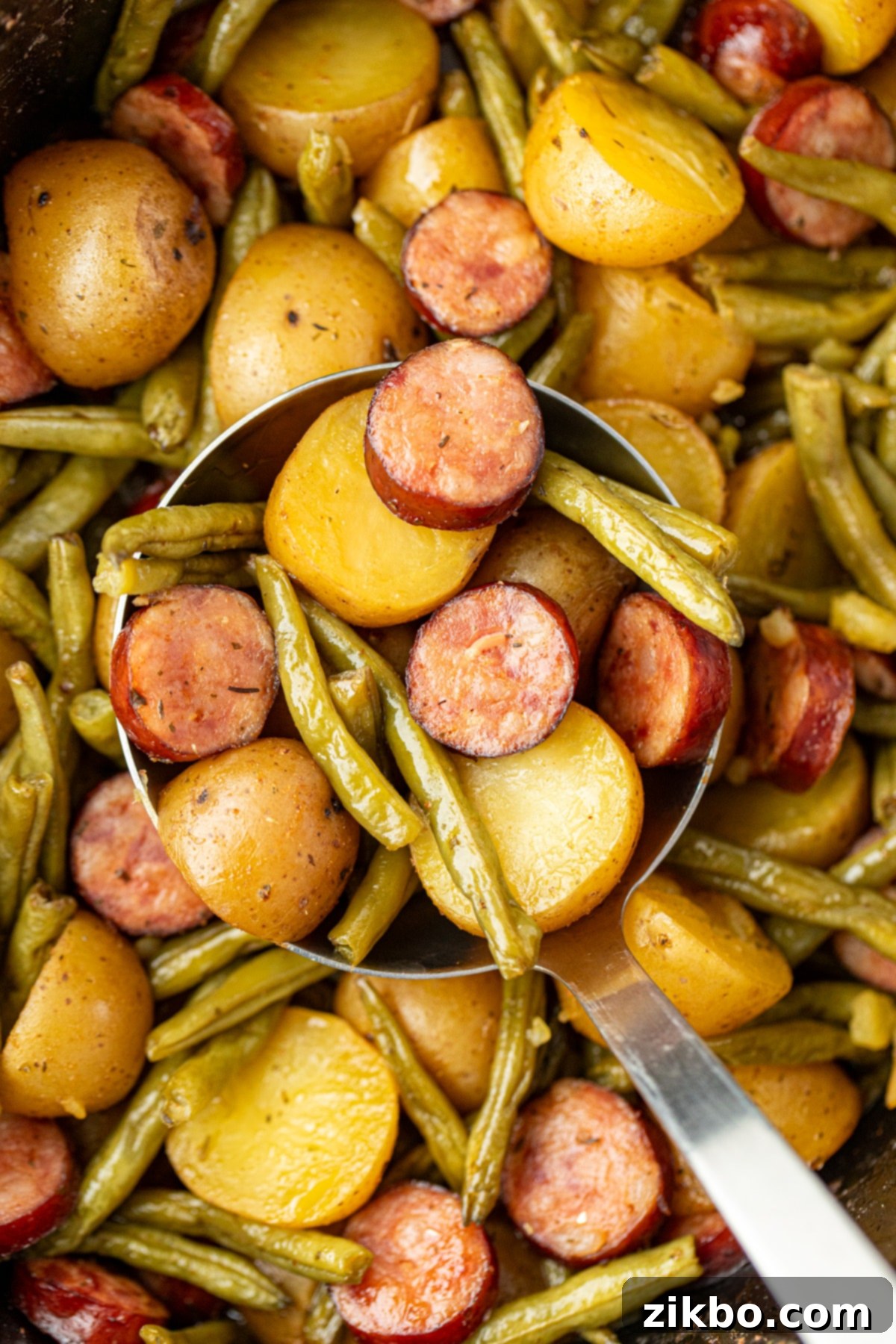 A large serving spoon lifting a generous portion of steaming, flavorful potatoes and sliced sausage from a slow cooker, ready for plates.