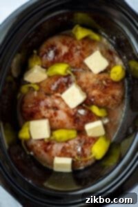 A slow cooker with chicken, seasoning, butter, and pepperoncini peppers.