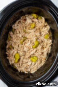 A slow cooker filled with juicy shredded chicken and a few pepperoncini peppers.