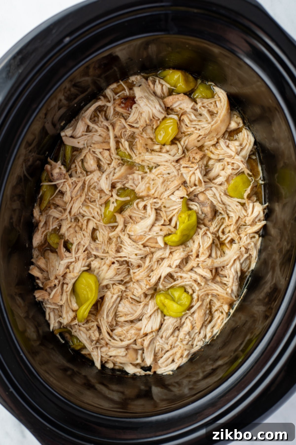 A slow cooker filled with juicy shredded chicken and a few pepperoncini peppers.