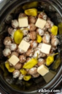 A slow cooker filled with meatballs, pepperoncini peppers, and butter slices.