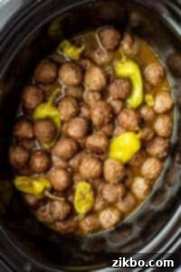 A slow cooker filled with cooked meatballs mixed with a thin sauce and pepperoncini peppers.