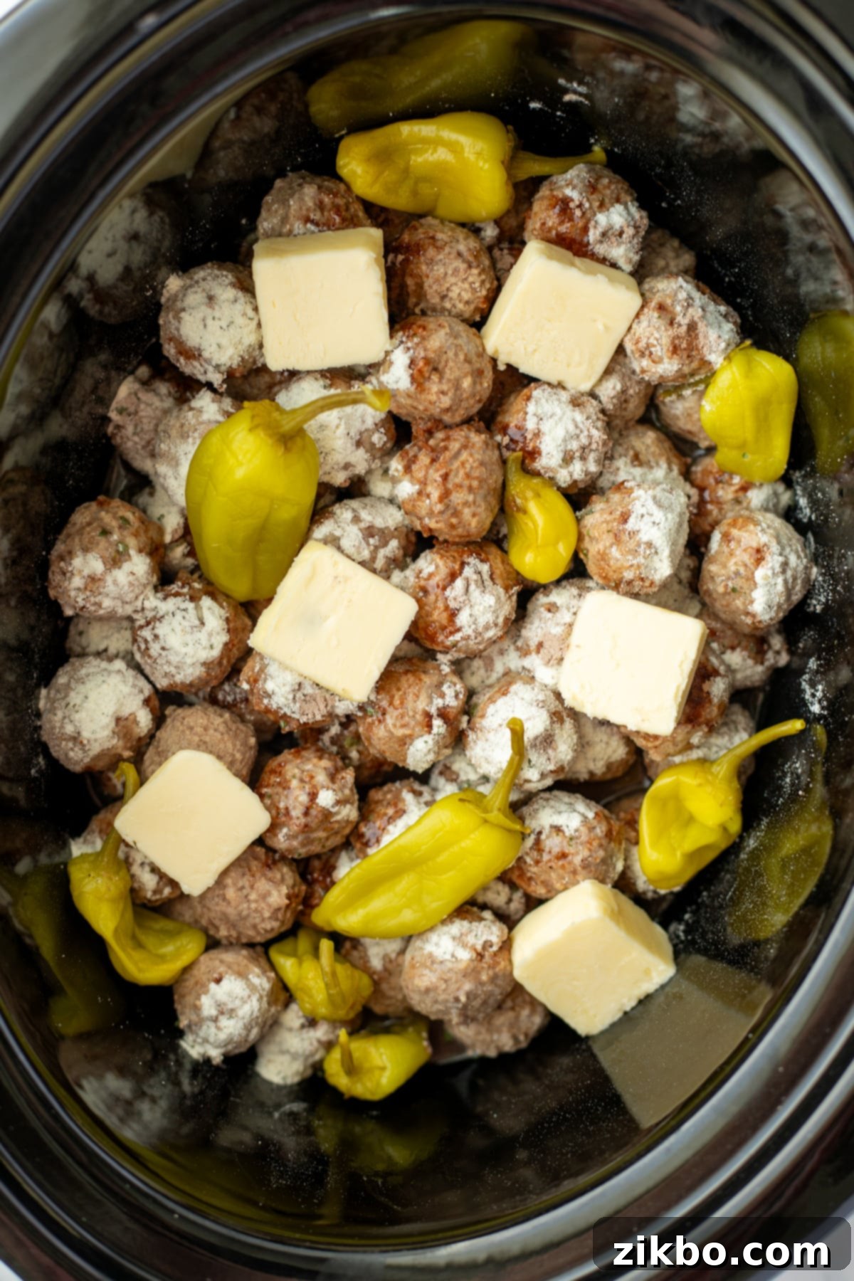 A slow cooker filled with meatballs, seasoned with ranch and au jus, topped with pepperoncini peppers and butter slices.