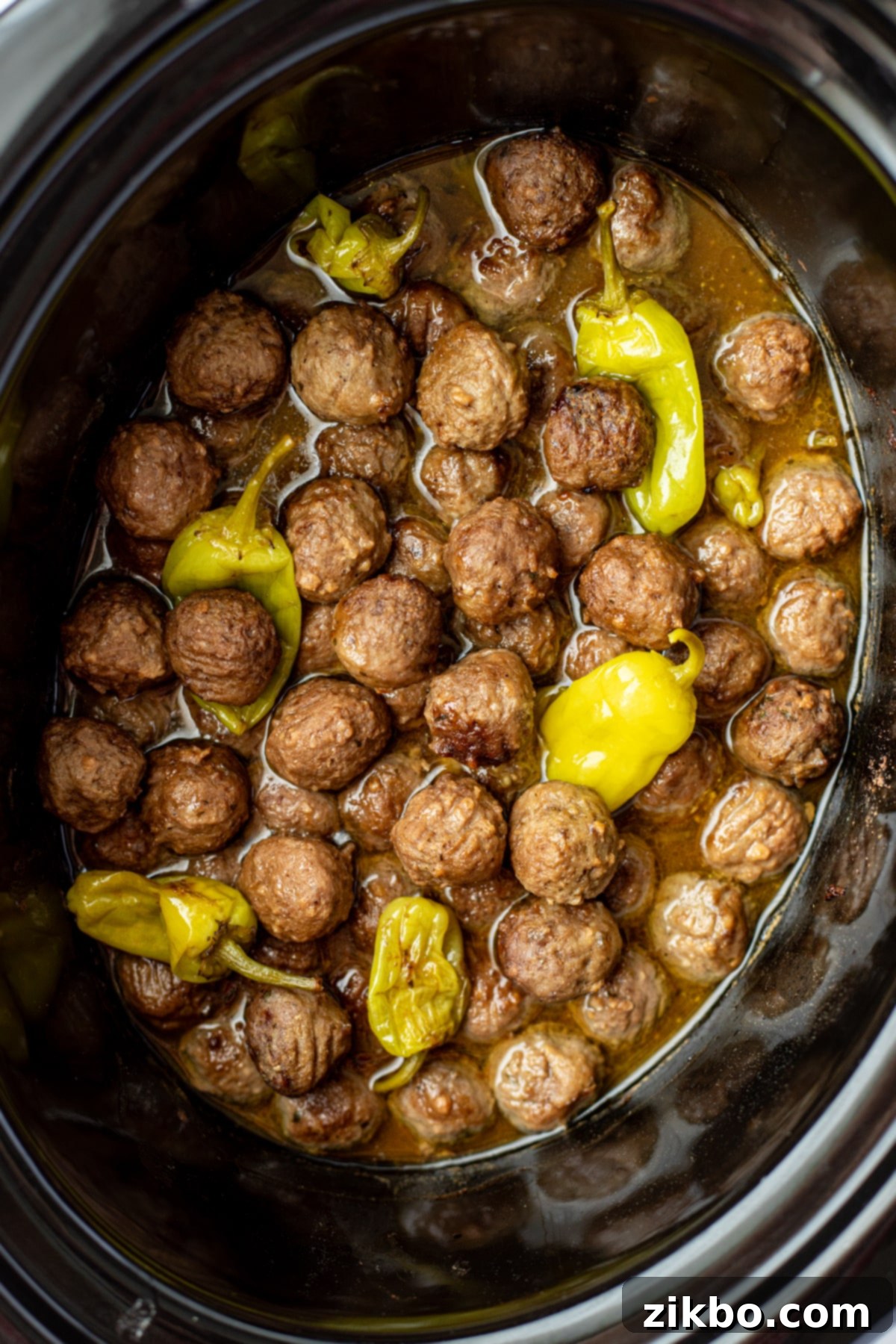 A slow cooker filled with cooked meatballs mixed with a thin, rich sauce and tender pepperoncini peppers.