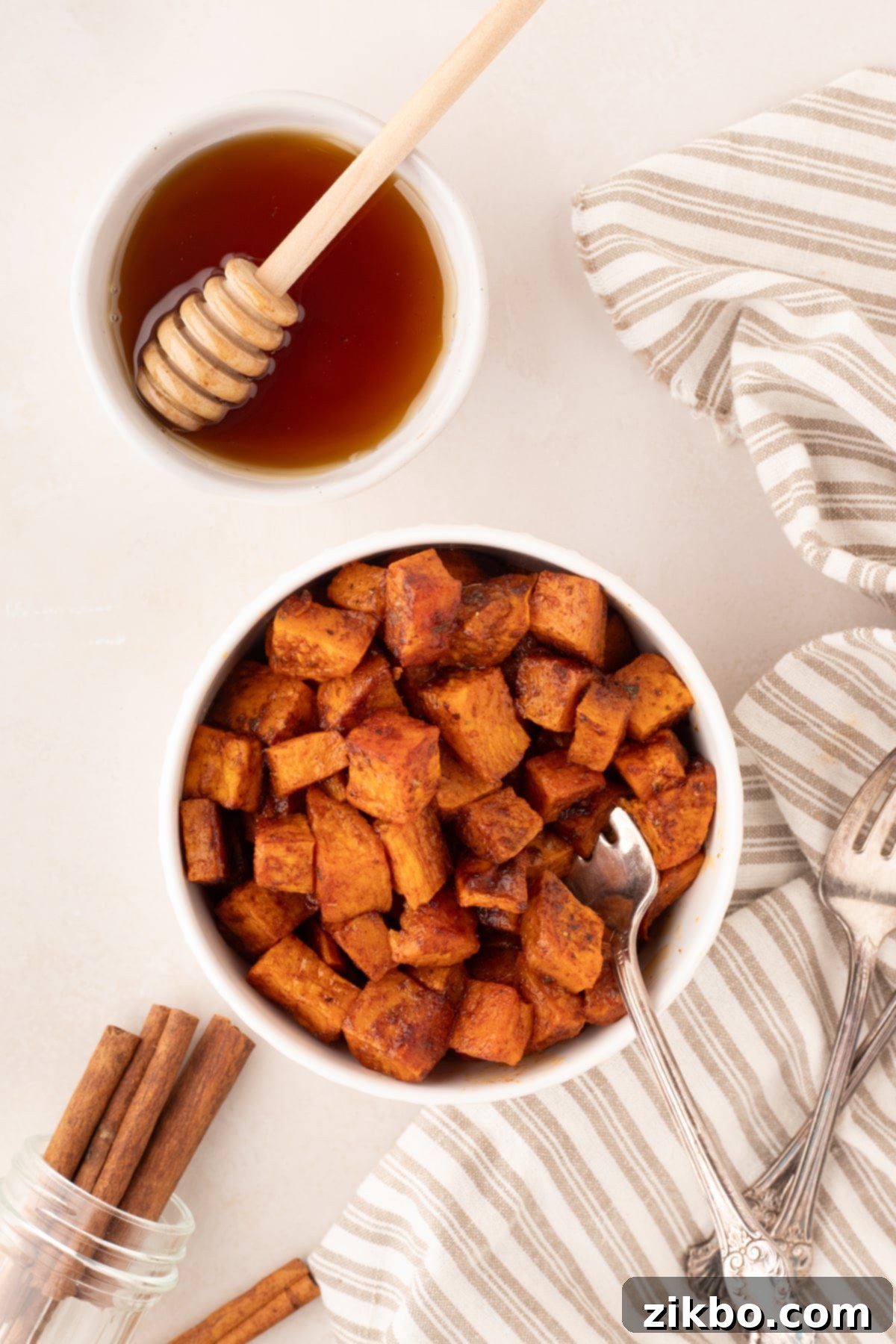 A bowl filled with golden-brown roasted sweet potatoes, with a small bowl of honey and a spoon on the side, ready for drizzling.