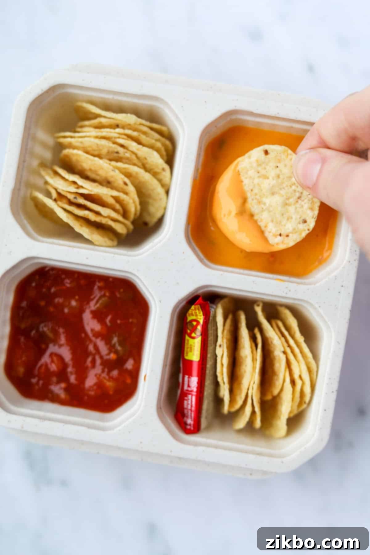 A close-up of a hand dipping a round tortilla chip into a container of warm queso cheese sauce, presented as part of a homemade nacho lunchable.