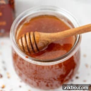 A beautiful jar of homemade hot honey with a loaded honey dipper, and scattered crushed red pepper flakes on a light colored counter. Perfect for any dish!