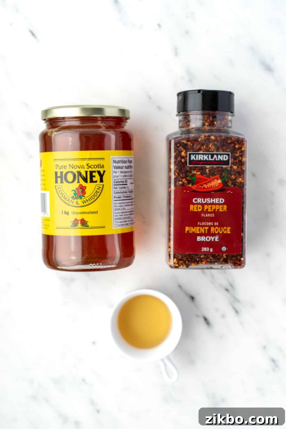 A jar of golden honey, a vibrant container of crushed red chili flakes, and a small bowl of amber apple cider vinegar, ready for making hot honey.