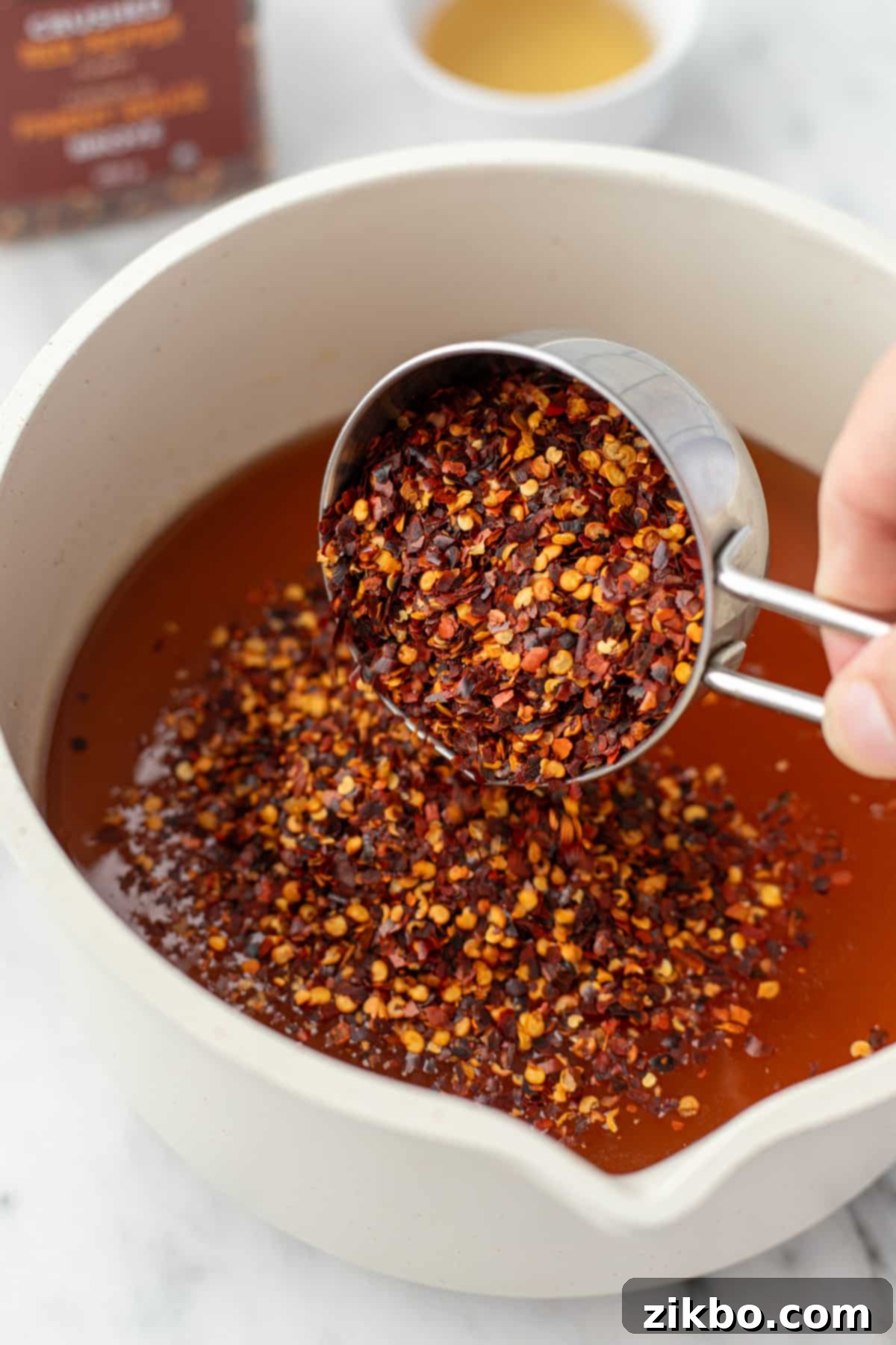 Adding a generous amount of dried red pepper flakes into a saucepan filled with warm, golden honey, ready for infusion.